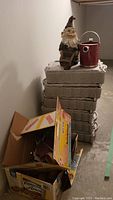 Stack of six rectangular gray seed trays and a small painted garden gnome statue alongside a red metal watering can.