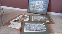 Four prints displayed including three framed prints with green mat board and gold frames and one unframed print with certificate of authenticity on floor against wall