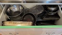 Drawer with metal mixing bowls, saucepans with lids, metal bakeware pan, wire cooling rack, and large metal strainer baskets.