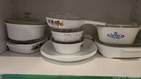 Shelf with stacked white casseroles featuring colorful fruit pattern and blue floral dish, some with clear glass lids.