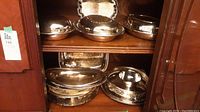 Two shelves filled with silver plated covered dishes and trays inside a wooden cabinet.