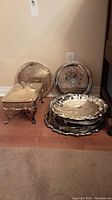 Photo showing seven round silver plate trays with engraved designs arranged on the floor and leaned against the wall with a rectangular silver plated food warmer beside them.