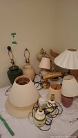 Overview of various ceramic and brass table lamps and several cream and beige lampshades on a table and floor.