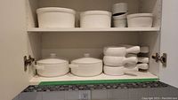 Two shelves inside a cabinet containing white ceramic dishes including covered casseroles and several white serving bowls and casseroles.