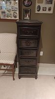 Front view of the tall dark wood cabinet showing six carved drawers and part of nearby chair and wall with framed pictures.
