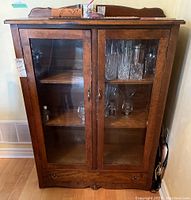 Closed view of the dark stained wood cabinet showing glass doors, wooden shelves inside with glassware, and a low drawer at the bottom