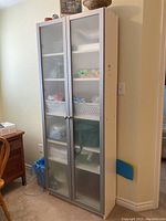 Front view of the tall white melamine cabinet with frosted plexiglass doors showing some contents inside.