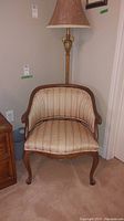 Front view of wood framed upholstered chair with curved arms and legs, displayed in a room corner beside a lamp and dresser.