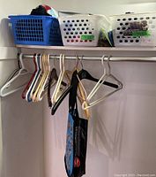 Closet rod with variety of plastic and wooden hangers, some with tags, baskets on top shelf containing assorted items including boxed light bulbs.