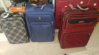 Three soft sided suitcases including black and white plaid, solid blue, and solid red, side by side on the floor.