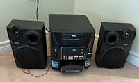 Full view of the RCA stereo system with two speakers positioned on the floor. The system is black and compact.