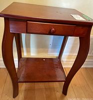 Front view of natural wood bow leg side table showing single drawer with round knob and bottom shelf with some scratches on surface.