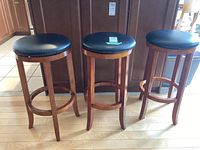 Set of three wooden swivel top stools with black cushioned seats arranged in a row on a wood floor in front of dark cabinetry.