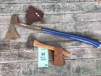 Both axe and hatchet shown together on a wooden surface with their leather covers.