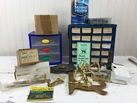 Setup of brass hardware collection including hooks, hinges, hose nozzle, two labeled drawer units and boxed kits.