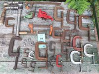 Top view of multiple clamps of various sizes arranged on a weathered wooden surface with some greenery in background, displaying many C-clamps and a pipe clamp.