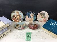 Five Edna Hibel collector plates displayed on a surface with four original boxes. Plates depict various mother and child groupings with delicate pastel colors and gold rims. The boxes show wear consistent with age.