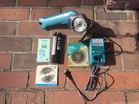 Photo showing Mikita cordless circular saw with one blade installed, two batteries (one inside saw, one beside it), a charger, and two extra saw blades in original packaging on brick surface.