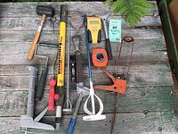 Overview of all tools on a weathered wooden surface showing a variety of hand tools for painting and caulking work including mallet, extension pole, caulk guns, scrapers, tape measure, and stud finder.