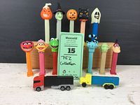 Photo showing the 15 PEZ dispensers arranged with 5 Halloween-themed heads, character heads from TV and movies, all empty, along with 2 tractor trailer truck toys in front.