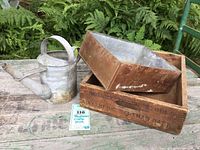 Wooden crate, metal box, and small galvanized watering can shown together outdoors on a weathered wooden surface with ferns in background.