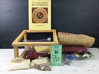 Photo showing the overall lot consisting of wooden frame, multiple bags of wool, and the rug hooking book arranged on a flat surface.