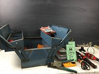 Photo showing the open blue metal toolbox with soldering irons, tools, and accessories inside on a white surface against a black background.