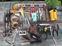 Photo of assorted used hand tools on wooden surface including clamps, adjustable wrench, Dremel rotary tool without bits, tape measures, hammer, pliers, and a flashlight/work light.