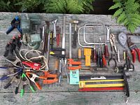 Wide view of all tools laid out on a weathered wooden bench outdoors, showing variety of hand tools including the vise, clamps, hand wrenches, hacksaw, screwdrivers, pliers, files, and the electric pencil engraver with power cord.
