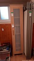 Front view of the tall narrow bookcase with frosted ribbed glass door showing the wooden frame and handle, placed beside a window and other household items.