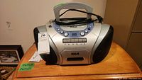 Front view of the RCA model ROD110B boombox showing the top handle, display, control buttons, speaker grilles on sides, and power cord wrapped around the handle.