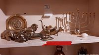 Full shelf view displaying five brass animal figurines, two brass plates, and a brass menorah candle holder, shows item condition and arrangement.