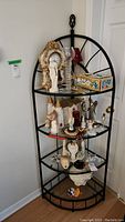 Full view of corner shelf with various decorative items on shelves. Black metal frame with four circular glass shelves and decorative metal finial on top.