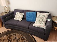 View of three-person dark upholstered couch with three pillows, curved front, and studded arms, situated on wooden floor near a decorative rug.