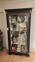 Full frontal view of tall black wooden display cabinet filled with various items. Shows ornate gold-red detailing on frame. Cabinet stands on flat feet with an overhanging crown molding.