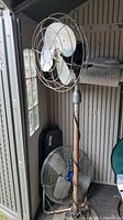 Tall industrial floor fan with metal blades and cage, visible on stand, beside smaller floor fan with plastic blades.