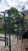 Black metal garden arch with curved top and decorative floral and tree panels shown in outdoor setting.