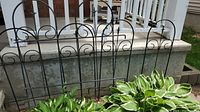 Three black metal garden trellises placed upright outdoors near flower bed and concrete steps with porch railing in background.