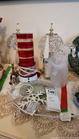 Wide view of lot including two ornate glass lamps with gold finials, a frosted glass snowman lamp holding a candle, a red and white striped glass decor piece, metal bell, and flower candles in green and red color.
