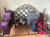 Six pieces of luggage placed on a metal bed frame base, showing variety of suitcase and duffel bag styles and colors.