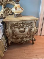 Front view showing one side table with detailed gold-tone floral and scrollwork carvings on three drawers. Light wood finish with curved drawer fronts and legs.