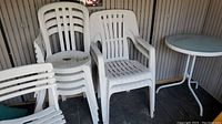 Six white plastic stacking patio chairs and two white folding plastic patio chairs arranged near a dark wooden floor and wall