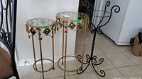 Three plant stands grouped together: two gold-toned with glass tops and decorative accents, one black-toned with scroll design.