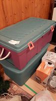 Two large plastic bins stacked, one with green lid and rope side handles, pink latches visible.