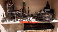 Shelf with silverplate plates, trays, goblet glasses, small bell, and other silver serving items showing tarnish and aged condition