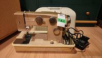 Full view of the White brand beige sewing machine sitting on its base, with attached cords and foot pedal. Beige carrying case behind.