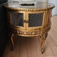 Front view of the wooden side table with gold finish, glass top, wire mesh double doors with floral knobs, and carved legs.