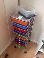 Tall metal shelving unit with perforated top and ten plastic drawers in various colors, standing on wheels in a corner against the wall.