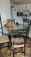 Full view of round glass table with black metal legs and four black metal chairs with padded tan seat cushions. Kitchen background visible.