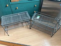 Two dark grey metal storage racks on a wood floor near a teal dresser and stainless steel refrigerator. The smaller rack has two mesh shelves and the larger rack has three mesh shelves.